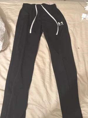 Svarta mjukisbyxor Under Armour - Snygga svarta mjukisbyxor från Under Armour med vit dragsko i midjan och logga på vänster lår. Byxorna har raka ben och är tillverkade i ett mjukt, stretchigt material som passar perfekt för chill eller träning.
