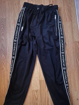 Svarta mjukisbyxor från Givenchy - Snygga svarta mjukisbyxor från Givenchy med vita loggor längs sidorna. Byxorna har resår i midjan med dragsko och dragkedjeförsedda fickor. Perfekta för en chill och trendig look. Materialet känns mjukt och följsamt.