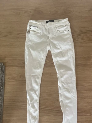 Vita skinny jeans med dragkedjedetalj - Säljer ett par vita skinny jeans med dragkedjedetalj vid fickan. Jeansen har klassisk femficksdesign och smal passform. Perfekta för dig som gillar en clean och stilren look. Materialet ser ut att vara bomull med lite stretch.