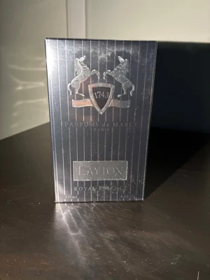 Parfums de Marly Layton 125ml - Exklusiv parfym från Parfums de Marly, modellen Layton. Flaskan rymmer 125 ml och kommer i en elegant mörkblå förpackning med silverdetaljer och emblem. Doften är en Eau de Parfum och förpackningen har en lyxig, stilren design med metallisk finish.