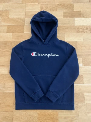Mörkblå Champion hoodie med huva - Snygg mörkblå hoodie från Champion med broderad logga på bröstet. Klassisk känguruficka framtill och stor huva. Perfekt för en avslappnad och sportig stil. Tillverkad i mjukt material som känns skönt mot huden.