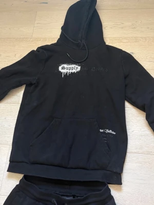 Svart set med hoodie och byxor - Säljer ett svart set med hoodie och matchande byxor. Hoodien har tryck med texten 'Supply' och en stor ficka framtill. Byxorna har dragsko i midjan och fickor på sidorna. Båda plaggen har en avslappnad passform och är perfekta för en chill stil.