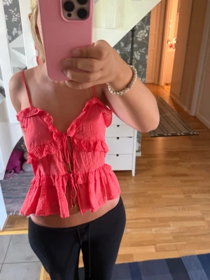 Volang topp med snörning - Säljer en söt rosa linnetopp med tunna axelband, volanger och snörning framtill. Toppen är croppad och har en luftig passform, perfekt för varma dagar. Materialet känns lätt och somrigt.