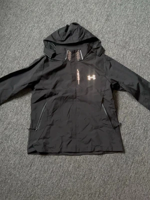 Svart vindjacka från Under Armour - Snygg svart vindjacka från Under Armour med huva och dragkedja framtill. Jackan har två sidofickor med dragkedja och reflekterande logga på bröstet. Tillverkad i lätt och tåligt material som står emot vind. Perfekt för aktiva dagar utomhus.
