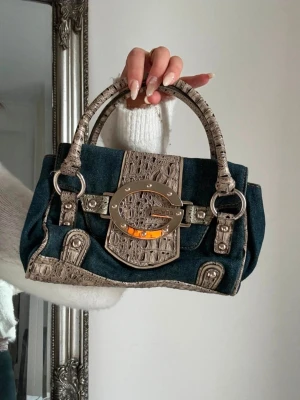Guess denim handväska med krokodetaj - Snygg handväska från Guess i mörkblå denim med beige krokodilpräglade detaljer och silverfärgad metallspänne framtill. Väskan har dubbla handtag och cool mix av material som ger en trendig look. Perfekt accessoar för att lyfta din outfit.