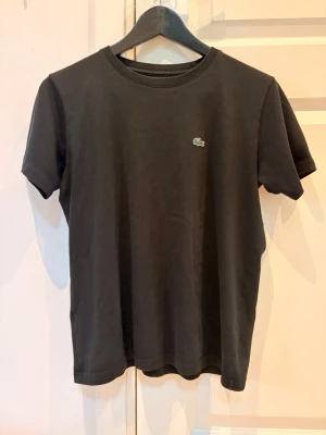 Svart t-shirt från Lacoste - Klassisk svart t-shirt från Lacoste med den ikoniska gröna krokodilloggan broderad på bröstet. T-shirten har rund halsringning och korta ärmar. Tillverkad i mjuk bomull för en bekväm känsla och enkel stil.