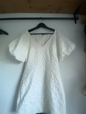 Vit kort klänning med puffärm - Säljer en vit kort klänning med v-ringning och stora puffärmar. Klänningen har ett diskret blommigt mönster i tyget och är tillverkad i ett strukturerat material som ger en lyxig känsla. Perfekt för dig som vill sticka ut med en unik och trendig look.