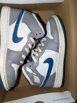 Nike Air Jordan 1 Mid grå/blå - Snygga Nike Air Jordan 1 Mid sneakers i vitt, grått och blått. Skorna har klassisk hög siluett, blå swoosh och detaljer samt vita skosnören. Tillverkade i läder med vadderad plös och yttersula i gummi. Perfekta för dig som gillar streetwear och ikonisk design.