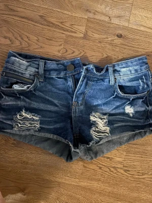 Blå slitna jeansshorts med dragkedja - Säljer ett par blå jeansshorts med slitningar. Shortsen har flera fickor, en extra dragkedja framtill och coola slitdetaljer både fram och bak. Materialet är klassisk denim och passformen är kort och tajt.