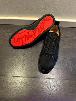 Svarta sneakers med röd sula - Stilrena svarta sneakers i mocka med klassisk låg profil och snörning. Skorna har en ikonisk röd sula med grafiskt mönster undertill. Insidan är beige och materialet ser exklusivt ut. Perfekta för dig som vill ha en clean och trendig look.