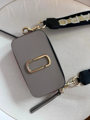 Beige axelväska med bred rem - Snygg Marc Jacobs beige axelväska i boxig modell med guldfärgad metallogo framtill. Väskan har en bred, justerbar axelrem med svart och guld textdetalj. Stängs med dragkedja och har dekorativa läderdetaljer. Perfekt för dig som gillar trendiga accessoarer.