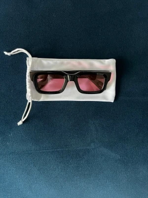 Svarta solglasögon med rosa glas - Säljer ett par trendiga solglasögon med svart, fyrkantig båge och rosa tonade glas. Kommer med en vit tygpåse för förvaring. Perfekta för dig som vill sticka ut med en cool accessoar i sommar.