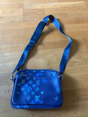 Blå axelväska - Snygg blå axelväska  med justerbar axelrem och ikoniskt monogrammönster i olika blå nyanser. Väskan har dragkedja upptill och innerfack för enkel organisering. Perfekt för dig som vill ha en trendig och praktisk accessoar.