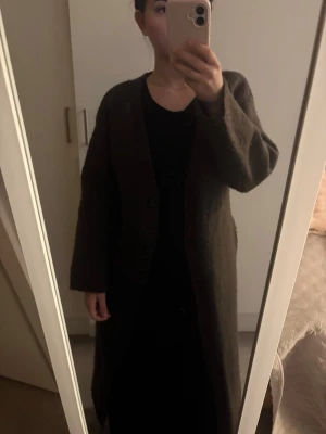 Lång brun cardigan med v-ringning - Mysig lång cardigan i mörkbrunt med v-ringad hals och raka ärmar. Jackan har en enkel knäppning framtill och är tillverkad i ett mjukt, stickat material som ger en avslappnad och trendig look. Perfekt att slänga över en outfit för extra värme.