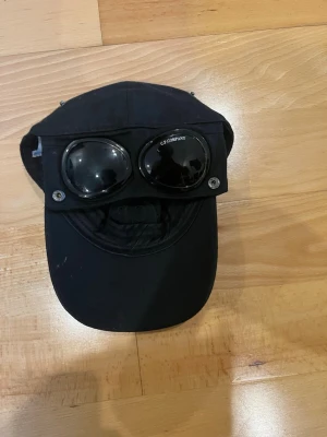 Svart keps med goggles från C.P. Company - Säljer en svart keps från C.P. Company med ikoniska inbyggda goggles framtill. Kepsen har böjd skärm, justerbar passform och är tillverkad i ett slitstarkt tyg. Perfekt för dig som vill sticka ut med en unik och streetinspirerad stil.