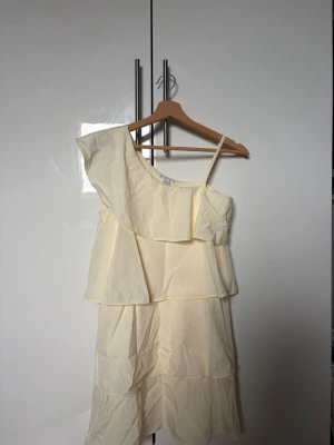 Ljusgul oneshoulder klänning volang - Säljer en söt ljusgul klänning med oneshoulder och volangdetaljer. Klänningen har lager på lager-effekt och ett axelband på ena sidan. Perfekt för sommarens alla tillfällen!