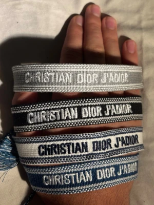 Christian Dior tygarmband - Fyra snygga tygarmband från Christian Dior med texten 'CHRISTIAN DIOR J'ADIOR' broderat i vitt, svart och blått. Armbanden har justerbara band med tofsar i matchande färger. Perfekt accessoar för att lyfta din outfit.