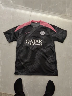 PSG svart matchtröja med rosa detaljer - Snygg svart PSG matchtröja från Nike med rosa axelpartier och logga. Tröjan har tryck med Qatar Airways på bröstet och Paris Saint-Germain klubbmärke. Perfekt för fotbollsträning eller att bära till vardags för att visa ditt lagstöd.