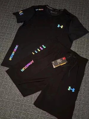 Svart träningsset från Under Armour - Säljer ett par svart träningsset från Under Armour med regnbågsfärgad logga och textdetaljer. Shortsen har snörning i midjan och är perfekta för träning eller chill. Materialet känns lätt och andas bra. Storlek S, helt ny 