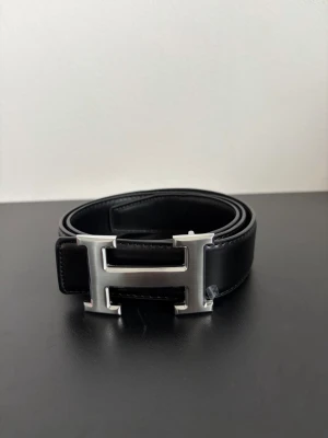Svart skärp med stor H-spänne - Snyggt svart skärp i skinn med ett stort, blankt H-format metallspänne. Perfekt accessoar för att lyfta vilken outfit som helst och ger en clean och stilren look. Klassisk design som funkar till både jeans och kostymbyxor.