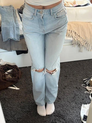 Ljusblå raka jeans med hål vid knäna - Säljer ett par ljusblå raka jeans med hög midja och slitna hål vid båda knäna. Jeansen har klassisk femficksdesign och stängs med knapp och dragkedja. 