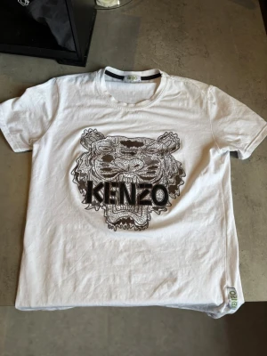 Vit Kenzo t-shirt med tigertryck - Säljer en vit t-shirt från Kenzo med ett stort, detaljerat tigertryck och logga på bröstet. Klassisk rund hals och korta ärmar. Tillverkad i mjuk bomull för skön känsla. Perfekt för dig som vill ha en statement-tee med ikonisk design. Passar mer som S.