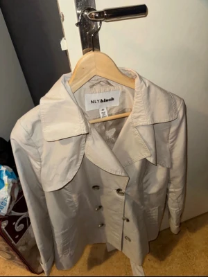 Beige trenchcoat från NLY Blush - Stilren beige trenchcoat från NLY Blush med dubbelknäppning och bred krage. Klassisk design med knappar framtill och bälte i midjan. Perfekt för dig som vill ha en tidlös och trendig jacka till garderoben.