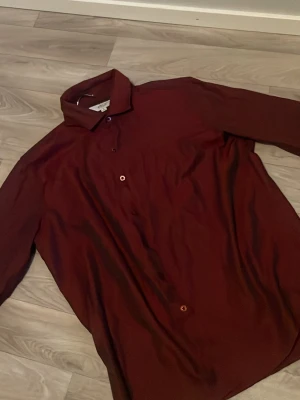 Mörkröd långärmad skjorta - Säljer en stilren mörkröd skjorta med klassisk krage och långa ärmar. Skjortan har knappar framtill och en rak passform. Perfekt för dig som vill ha en enkel men snygg skjorta i garderoben.