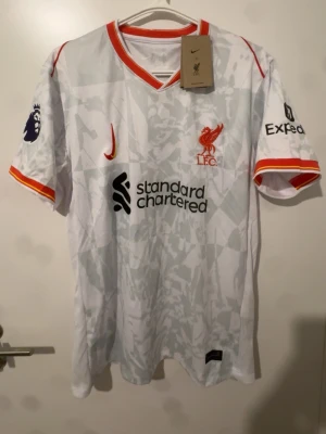 Liverpool bortatröja Nike vit/röd - Liverpool FC bortatröja från Nike i vitt med ljusgrått mönster och röda detaljer. Tryck med klubbmärke, sponsorloggor och Premier League-märke på ärmen. Nummer 11 och M. Salah på ryggen. Tillverkad i lätt och ventilerande material.