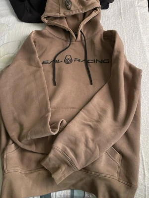 Beige hoodie från Sail Racing - Säljer en beige hoodie från Sail Racing med svart logga och text på bröstet. Hoodien har huva med dragsko, magficka och ribbade muddar vid ärmar och nederkant. Perfekt för en avslappnad och stilren look.