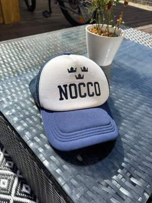 NOCCO keps - Snygg keps från NOCCO i blått och vitt med tre kronor-tryck framtill. Framsidan är vit med svart text och symbol, medan skärmen och baksidan är blå. Klassisk trucker-modell med mesh på sidorna och justerbar passform.