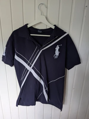 Svart kortärmad t-shirt med vita ränder - Snygg svart t-shirt med korta ärmar och vita diagonala ränder framtill. Stor logga med polospelare på bröstet och siffror på ena ärmen. Skön krage och avslappnad passform, perfekt till jeans eller shorts.