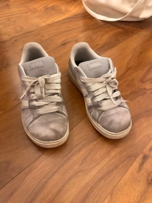 Grå Adidas sneakers med snörning - Snygga grå sneakers från Adidas med klassiska tre ränder på sidan. Skorna har en låg profil, rund tå och breda snören. Ovandelen är i mocka och insidan är fodrad med textil. Inga hål endast missfärgningar som syns på bilden 