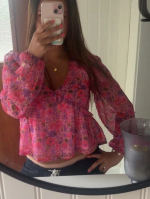 Blommig rosa blus med v-ringning - Säljer en söt blommig blus i rosa och lila toner med v-ringning och volang nedtill. Blusen har långa, transparenta ärmar och ett luftigt, flowy material som känns lätt och somrig. Perfekt för dig som gillar färg och mönster!