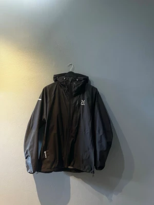 Svart vindjacka från Haglöfs - Snygg svart vindjacka från Haglöfs med huva och dragkedja framtill. Jackan har minimalistisk design, diskret logga på bröstet och är tillverkad i ett lätt, vattenavvisande material. Perfekt för blåsiga dagar och outdoor-äventyr.