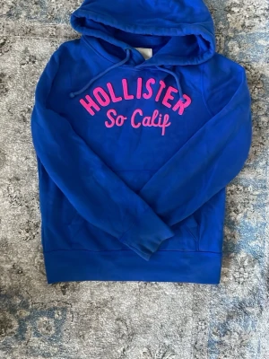 Blå hoodie från Hollister med tryck - Snygg blå hoodie från Hollister med huva och dragsko. Framtill står det 'Hollister So Calif' i rosa text. Hoodien har en klassisk känguruficka och är gjord i mjukt material, perfekt för chill dagar.