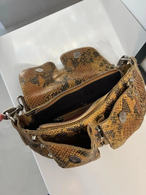 Brun handväska i ormmönstrat skinn - Cool handväska i brunt ormmönstrat skinn med flera fickor framtill och silverfärgade nitar. Väskan har bred axelrem med detaljer och magnetknappar på fickorna. 