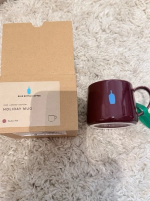 Holiday Mug från Blue Bottle Coffee - Säljer en limited edition Holiday Mug från Blue Bottle Coffee i en djup röd färg med blå logga. Muggen är cylindrisk och tillverkad i keramik. Perfekt för kaffeälskare som vill ha något unikt i köksskåpet.
