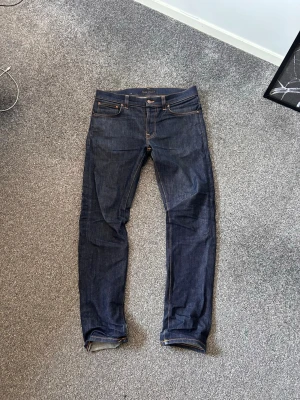 Mörkblå Nudie Jeans men fades på gång - Snygga mörkblå jeans i klassisk modell med raka ben och fem fickor. Jeansen har kontrastsömmar och detaljer på bakfickorna. Tillverkade i kraftigt denimtyg som ger en tidlös look. Perfekta för dig som gillar stilrena och enkla jeans. 