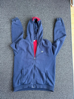 Blå zip hoodie från Lacoste - Snygg blå hoodie med dragkedja från Lacoste. Hoodien har en röd rand med Lacoste-tryck på ryggen och röd insida i huvan. Klassisk krokodil-logga på bröstet och två fickor framtill. Perfekt för en chill och sportig look.