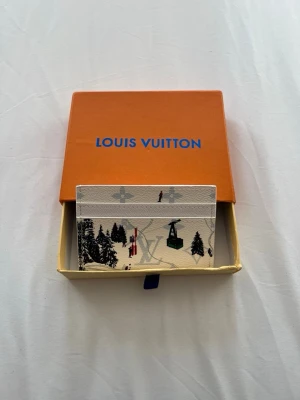Louis Vuitton vit korthållare med vintermotiv - Vit korthållare från Louis Vuitton med vinterinspirerat tryck som visar skidåkare, liftar och granar. Klassisk monogramstruktur i bakgrunden och flera fack för kort. Kommer i original orange Louis Vuitton-box.