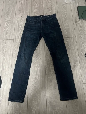 Mörkblå jeans från BOSS - Snygga mörkblå jeans från BOSS med rak passform och klassisk femficksdesign. Jeansen har diskreta slitningar och en stilren läderpatch med BOSS-logga bak i midjan. Perfekta till en avslappnad men ändå stilren look.