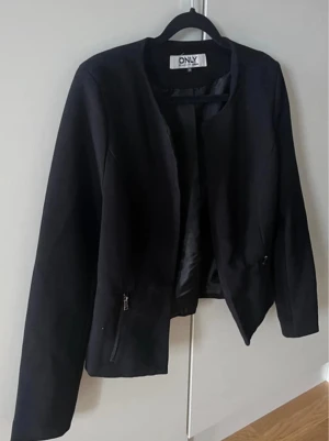 Svart blazer från ONLY - Stilren svart blazer från ONLY med rundad krage och dragkedjor vid fickorna. Kavajen har en rak passform och är lätt fodrad för extra komfort. Perfekt för dig som vill ha en enkel men snygg look.