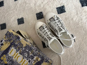 Gråvita Dior sneakers med mönster - Säljer ett par gråvita Dior sneakers med klassiskt monogrammönster och vita snören. Skorna har vit sula med stjärndetalj undertill och är låga i modellen. Materialet är canvas med gummidetaljer. Perfekta för dig som gillar exklusiv streetstyle.