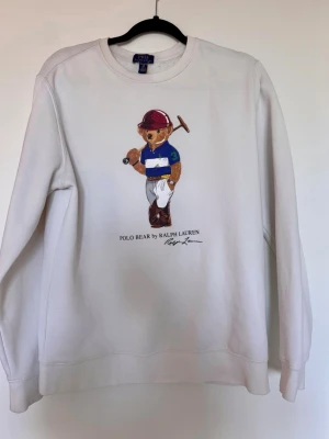 Vit stickad tröja Polo Bear - Vit stickad tröja från Polo Ralph Lauren med det klassiska Polo Bear-motivet framtill. Tröjan har rund halsringning, långa ärmar och ribbade muddar. Materialet ser ut att vara mjukt och bekvämt, perfekt för en avslappnad stil. Strl 16-18 år motsvarar 170 eller S herr
