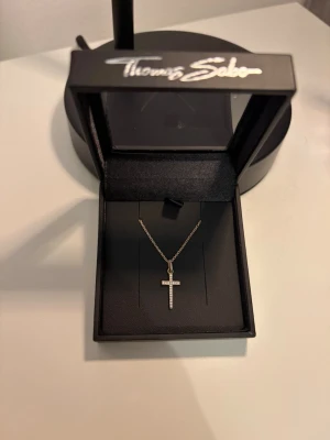Thomas Sabo halsband med kors - Stilrent halsband från Thomas Sabo med ett korsformat hänge dekorerat med små gnistrande pärlor. Kedjan är tunn och silverfärgad, och halsbandet ligger i originalask. 