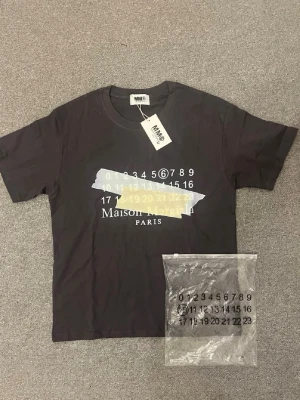  Maison Margiela t-shirt - Cool svart t-shirt från Maison Margiela med tryck av siffror och text på bröstet. T-shirten har korta ärmar och rund halsringning. Perfekt för dig som gillar streetwear och vill sticka ut med en unik design.