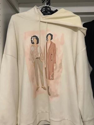 Vit hoodie med grafiskt tryck - Säljer en oversized vit hoodie med huva och dragsko. På framsidan finns ett grafiskt tryck av två stilrena figurer i pastellfärger. Hoodien har långa ärmar och mjukt material, perfekt för en chill och trendig look.