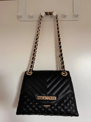 Svart quiltad axelväska Steve Madden - Snygg svart axelväska från Steve Madden med quiltat mönster och guldfärgade detaljer. Väskan har en kedjerem med inslag av svart material och ett stort guldfärgat logotypmärke framtill. Perfekt för dig som gillar trendiga accessoarer.