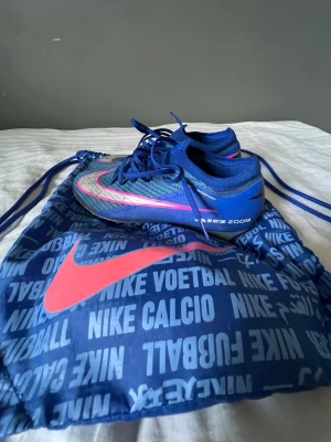 Nike blåa fotbollsskor Zoom - Snygga blåa Nike fotbollsskor med rosa och silver detaljer samt Zoom-teknologi. Skorna har låg profil och snörning för bra passform. Ovandelen är i textil och syntetmaterial. Perfekta för dig som vill sticka ut på planen.